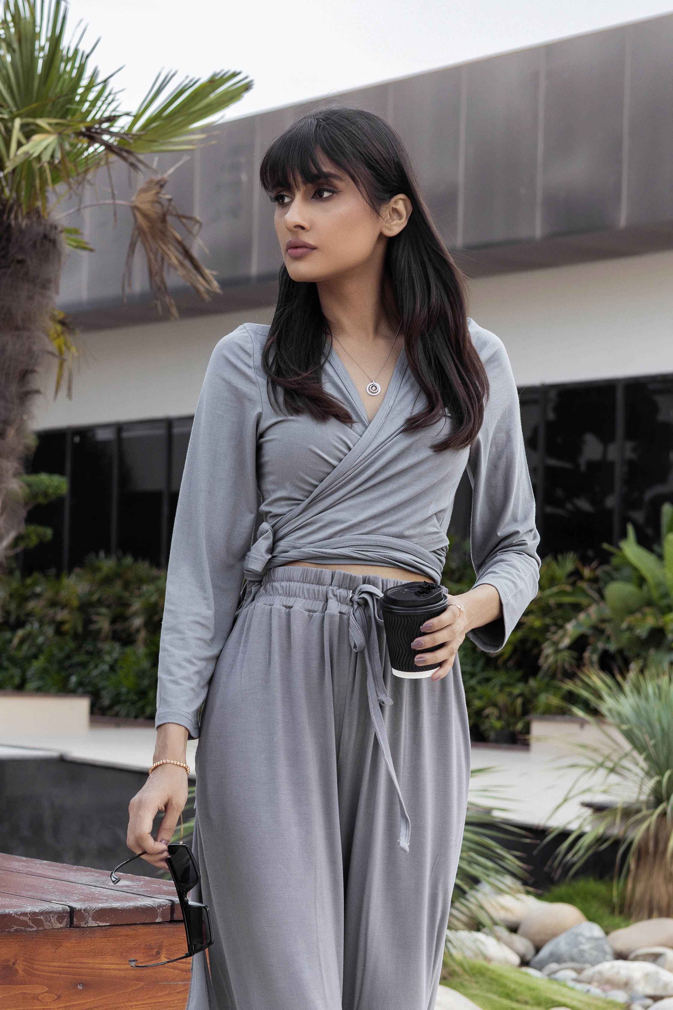 Woman in a gray outfit holding a coffee cup outdoors with plants and a building in the background