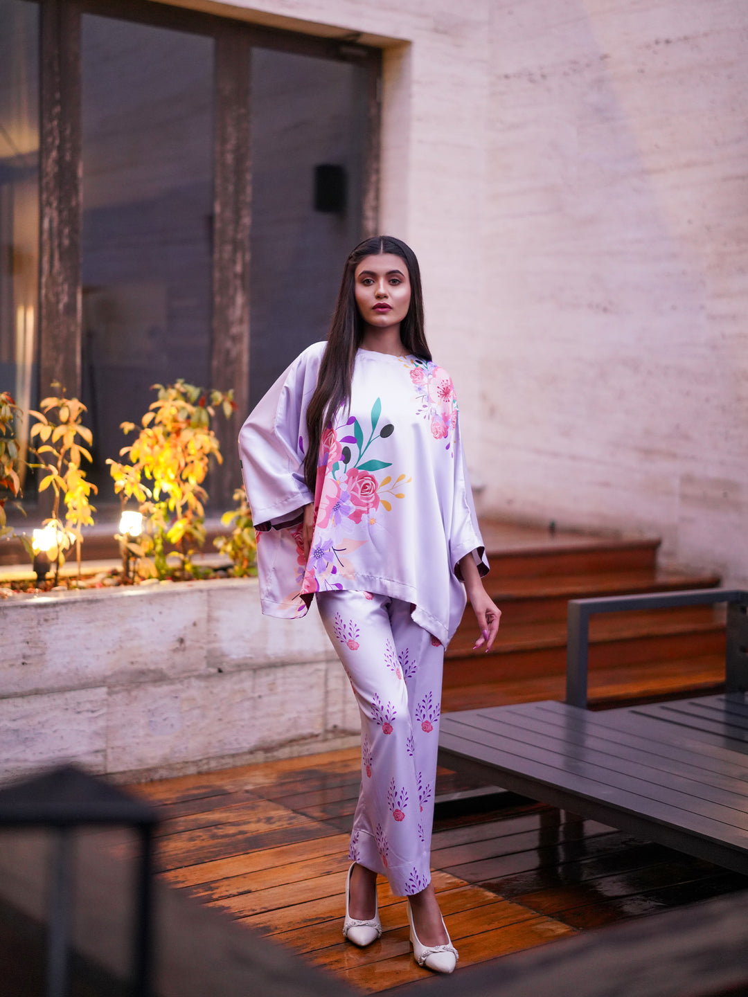 A woman wearing a white and pink floral printed kaftan top with a boat shape neckline and straight matching pants, standing near a window with plants.