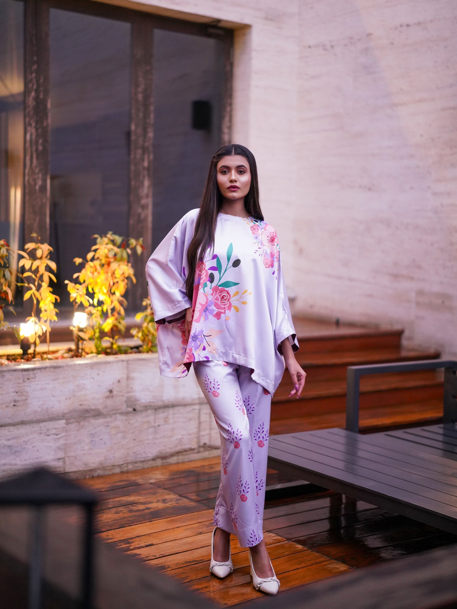 A woman wearing a white and pink floral printed kaftan top with a boat shape neckline and straight matching pants, standing near a window with plants.
