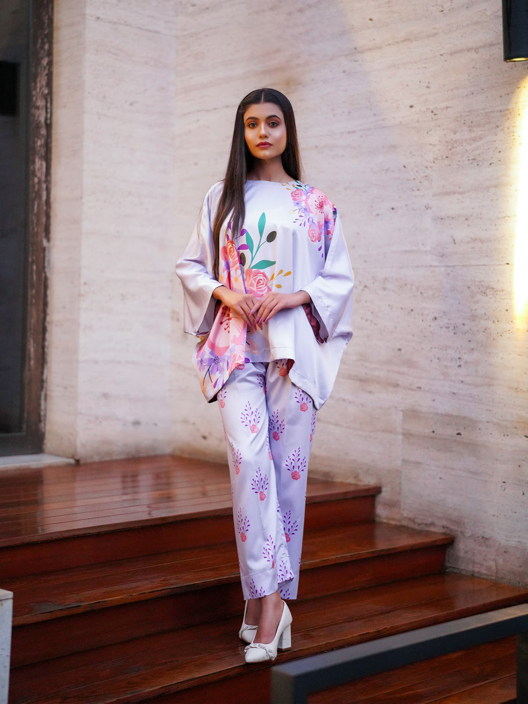 A woman standing on a staircase wearing a traditional, floral print kaftan top with a boat neckline and matching trousers, with a romantic and elegant pose.