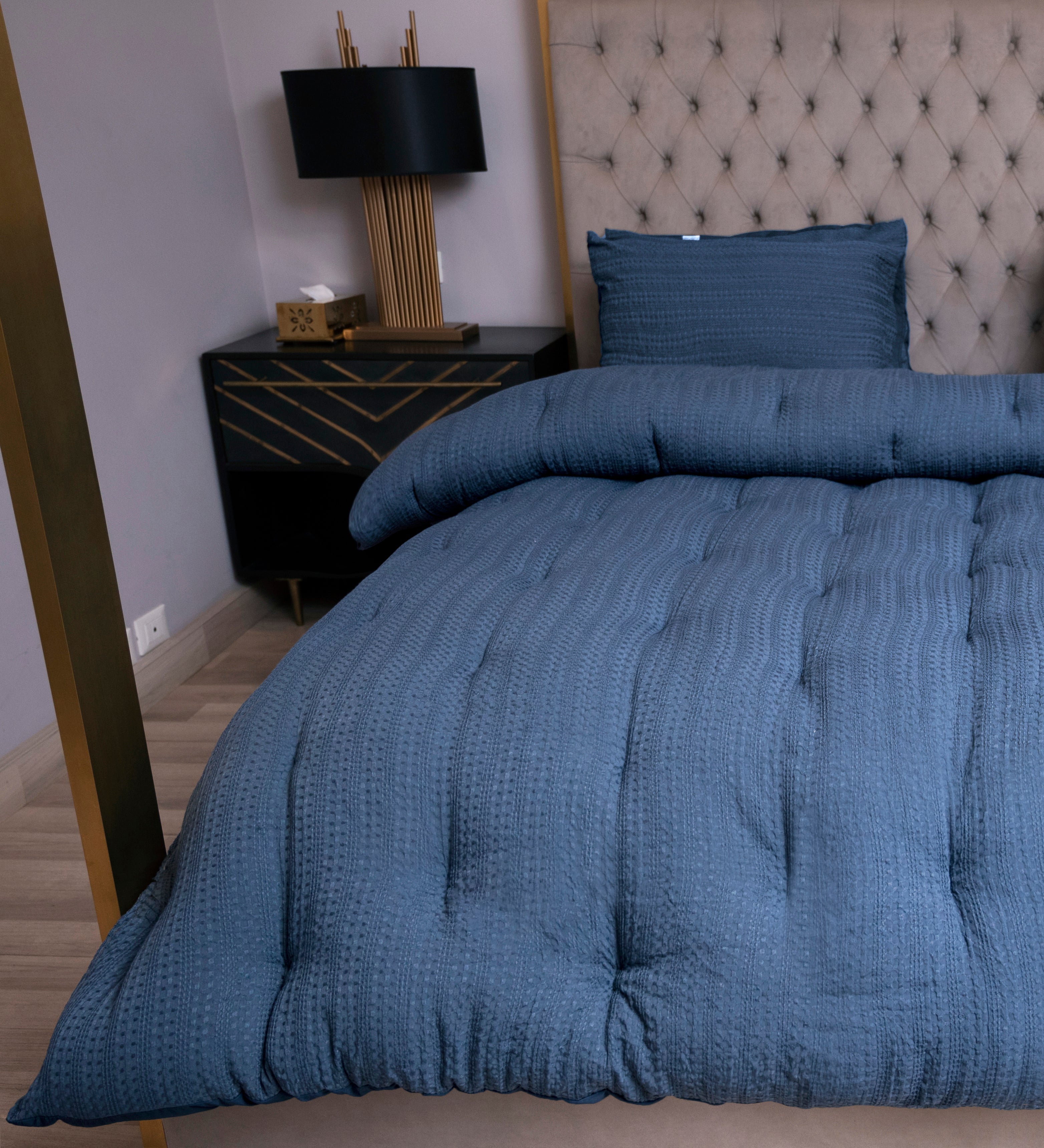 Blue textured comforter on a bed with a nightstand and lamp in the background.